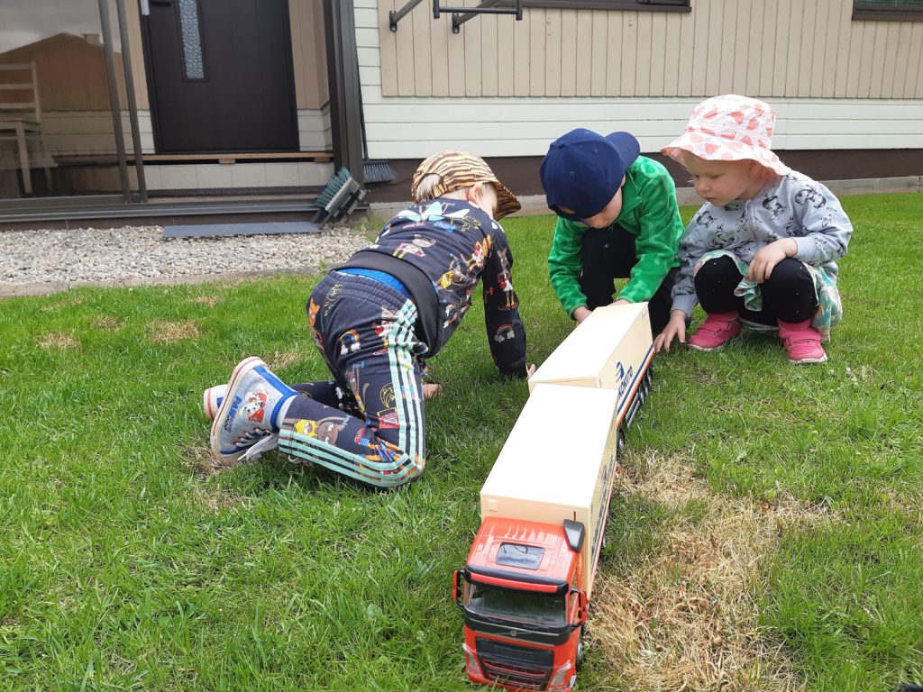 Kolme lasta leikkii pihanurmikolla leikkikuorma-autolla.