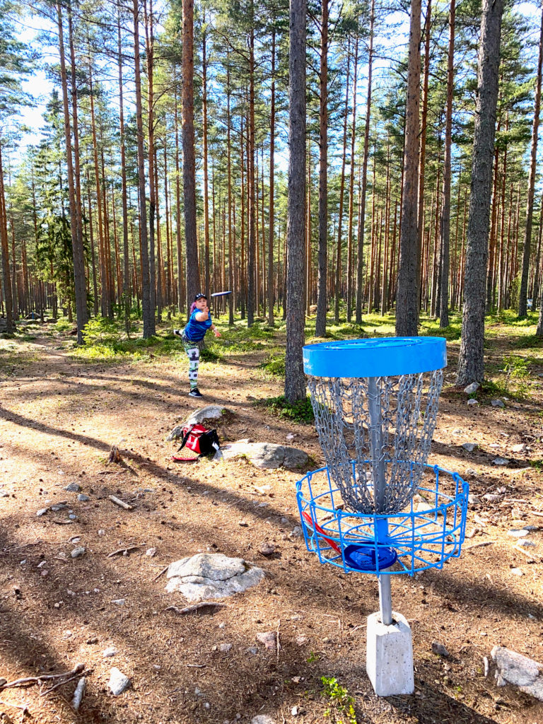 Metsämaisema, jossa etualalla frisbeegolf -kori, jota kohti pelaaja heittää kiekkoa