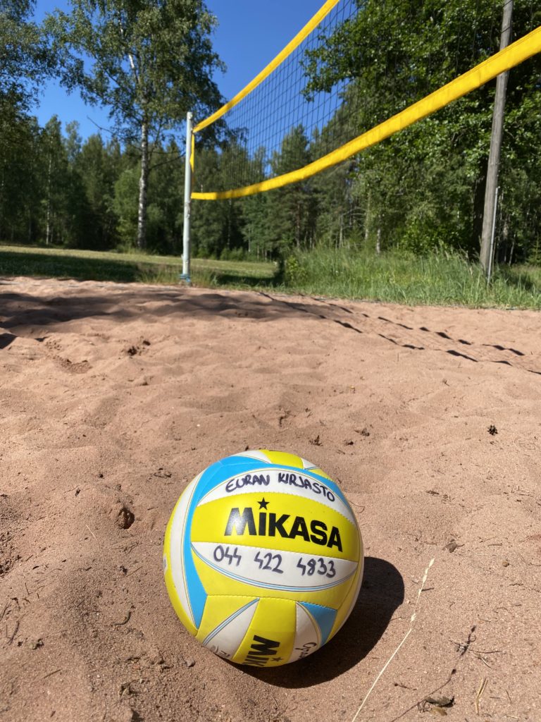 Kuvassa kesäinen rantalentis kenttä, jossa etualalla pallo ja taustalla hiekkakenttä ja verkko